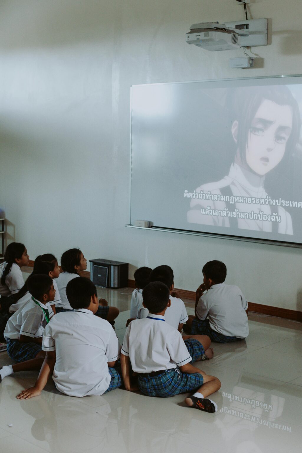Group of school children watching anime on a projector screen indoors.