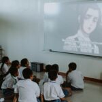 Group of school children watching anime on a projector screen indoors.